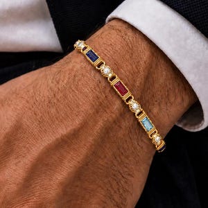 Op de afbeelding: Gouden armband met afwisselende rechthoekige edelstenen in blauw, rood en lichtblauw, afgewisseld met kleine diamanten. De armband wordt om een pols gedragen, met een donker pak en witte manchet op de achtergrond.