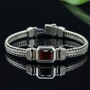 May include: A silver bracelet with a woven chain design and a rectangular, deep red gemstone at the center. The bracelet has a secure clasp and decorative geometric patterns. The jewelry is displayed on a reflective black surface.