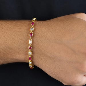 May include: A gold-tone chain bracelet featuring alternating round and rectangular-cut red gemstones and clear white stones. The bracelet is composed of a series of linked settings that hold the stones in a repeating pattern along the wrist.