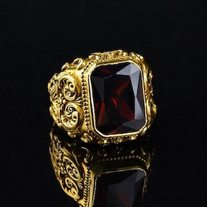 Può includere: Un anello color oro con un design ornato e una grande pietra preziosa rettangolare rosso scuro. La fascia dell'anello presenta dettagliate volute ed è posta su uno sfondo nero, creando un contrasto sorprendente.