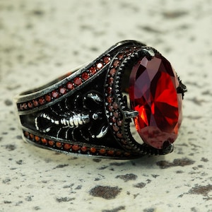 May include: A silver ring with a large, oval-cut, deep red gemstone as the centerpiece. The ring features a detailed scorpion design on the band and is accented with smaller red stones. The setting is dark, contrasting with the bright red of the main stone.