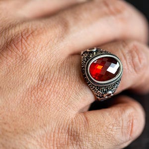 Peut inclure: Une bague en argent ornée d'une pierre précieuse rouge ovale et facettée. La bague présente un motif orné et détaillé autour de la pierre précieuse et sur le corps. La bague est portée au doigt.