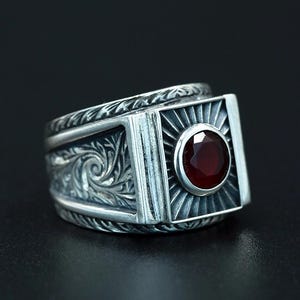 May include: A silver ring with a square face featuring a radiant design around a round, deep red gemstone. The band is detailed with ornate, swirling patterns. The ring is set against a dark background.