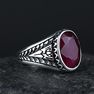 Op de afbeelding: Een zilveren ring met een ovale, facetgeslepen, dieprode edelsteen. De ringband heeft een sierlijk, gegraveerd ontwerp met blad- en geometrische patronen. De edelsteen is gezet in een gepolijste zilveren zetting.