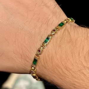 May include: Gold bracelet featuring rectangular green and round red gemstones. The bracelet has an ornate design with detailed metalwork. The gemstones are set in a repeating pattern along the length of the bracelet.
