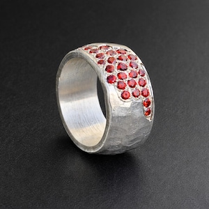 May include: A silver ring with a textured band and a cluster of small, round, red gemstones. The gemstones are set in a curved pattern across the top of the ring, creating a sparkling accent.