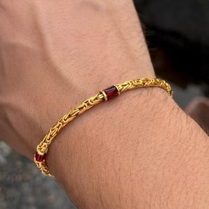 May include: A gold-colored bracelet with a chain-like design. The bracelet features three rectangular, deep red gemstones. The bracelet is worn on a wrist, showcasing its intricate details and warm tones.