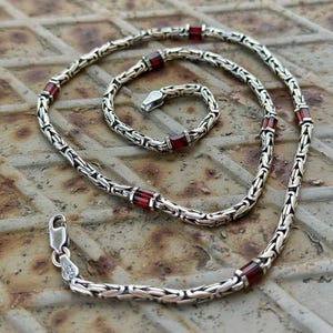 Può includere: Un set di collana e bracciale in argento con pietre rosse rettangolari. I gioielli presentano un design a catena dettagliato e una chiusura a fermaglio sulla collana. Il set è esposto su una superficie metallica strutturata.