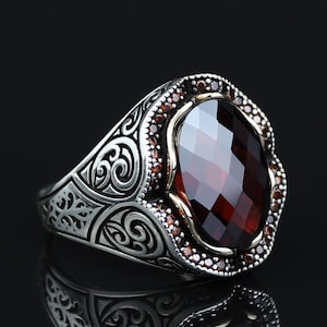 Peut inclure: Une bague en argent ornée d'une grande pierre précieuse facettée rouge foncé de forme ovale au centre. Le sertissage est entouré d'une bordure de petites pierres rouge foncé et de motifs de volutes gravés sur les côtés de l'anneau.