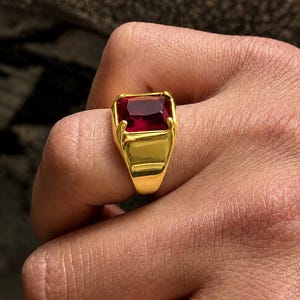 May include: A gold ring featuring a large, rectangular, deep red gemstone. The ring has a thick, polished gold band and a setting that securely holds the gemstone. The design is classic and bold, suitable for various occasions.