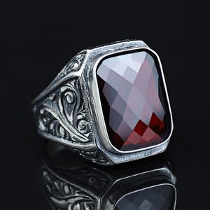 Pode incluir: Um anel de prata com uma grande pedra preciosa retangular facetada de cor vermelho escuro. O anel tem um design ornamentado e gravado na banda. O anel é exibido em uma superfície preta reflexiva.