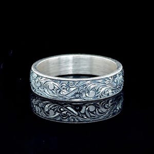 Könnte beinhalten: Ein silberfarbener Bandring mit einem komplizierten, tief eingravierten Blumen- und Rankenmuster auf der gesamten Außenfläche. Das Metall hat eine polierte Oberfläche mit abgedunkelten vertieften Bereichen, die das Relief betonen.