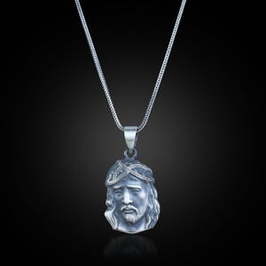 May include: A silver pendant necklace featuring a detailed depiction of a religious figure's face. The pendant is suspended from a delicate chain. The figure has a beard and a crown-like design on the head. The background is black.