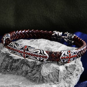 May include: A brown leather bracelet with a silver clasp and two silver decorative pieces with red gemstones. The bracelet is woven and has a braided design.