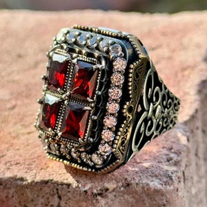May include: A silver ring with a large square red gemstone surrounded by smaller white gemstones. The ring has an intricate design with a black background.