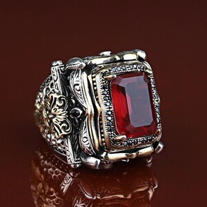 Puede incluir: Un anillo de plata con una gran gema roja rectangular. El anillo tiene detalles intrincados con acentos dorados y pequeñas piedras negras. El diseño es ornamentado y detallado, adecuado para una pieza llamativa.