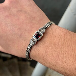 May include: A silver bracelet with a rectangular, deep red gemstone at the center. The bracelet features a braided chain design with decorative square accents on either side of the gemstone. The bracelet is worn on a wrist.