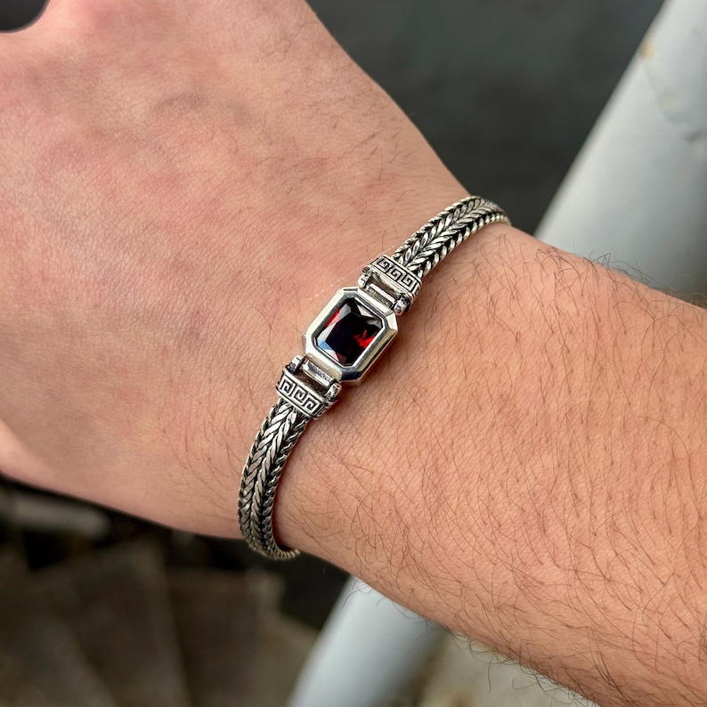 May include: A silver bracelet with a rectangular, deep red gemstone at the center. The bracelet features a braided chain design with decorative square accents on either side of the gemstone. The bracelet is worn on a wrist.