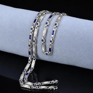 Puede incluir: Un collar plateado con piedras preciosas azules rectangulares y redondas. El collar está colocado sobre una tela azul claro, mostrando su intrincado diseño y detalles brillantes. El cierre es visible en la parte inferior.