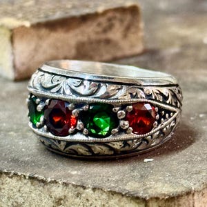 May include: A silver ring with an intricate floral design and three gemstones. The gemstones are red and green.