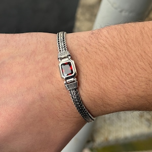 May include: Silver bracelet featuring a rectangular, deep red gemstone at the center. The bracelet has a woven chain design with decorative square accents on either side of the gemstone. The bracelet is worn on a wrist.