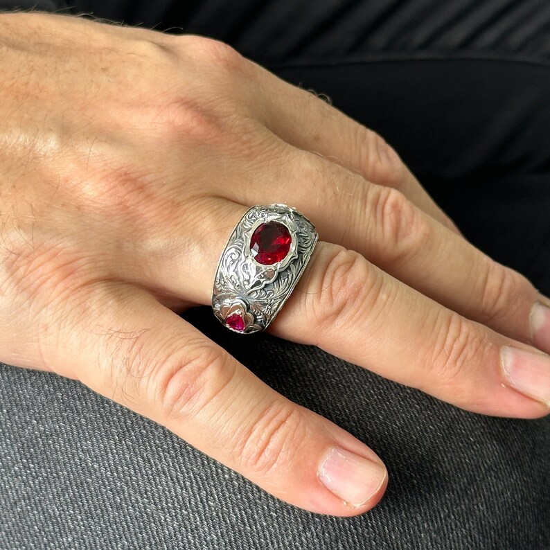 Peut inclure: Une bague en argent avec un design complexe et trois pierres pr&eacute;cieuses rouges. La bague est port&eacute;e au doigt d'une personne.