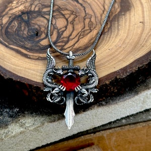 Puede incluir: Collar con colgante de plata con una espada con una piedra preciosa roja, flanqueada por dos dragones con las alas extendidas.
