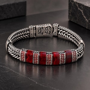 Könnte beinhalten: Ein silbernes Armband mit geflochtenem Kettenmuster und einem rechteckigen Kastenschloss. Der Mittelteil zeigt drei glatte, tiefrote rechteckige Steine, die sich mit kleineren, texturierten silbernen Zwischenstücken und gemusterten Gliedern abwechseln.
