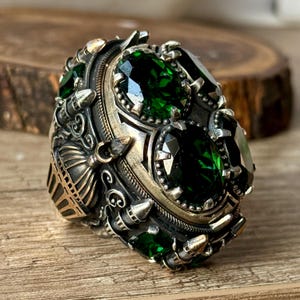 May include: A silver ring with intricate details and three large green gemstones. The ring has a gold-colored band and a design that resembles a dome or a building.