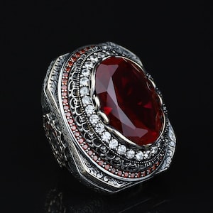 May include: A silver ring featuring a large oval-cut deep red centre stone, surrounded by a halo of small clear white stones and an outer border of tiny red stones. The metal band displays intricate engraved patterns along the sides and base.
