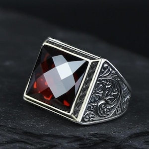 May include: A silver ring with a large, square, faceted, deep red gemstone. The ring has an ornate, engraved design on the sides. The gemstone is set in a silver bezel. The ring is displayed against a dark background.