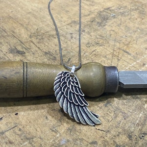 Puede incluir: Un collar de cadena de plata con un colgante de ala de plata. El ala está detallada con plumas texturizadas.