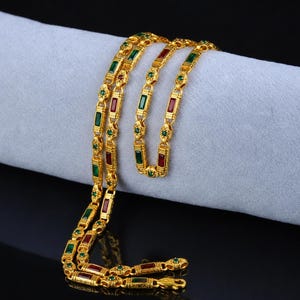 May include: A gold-colored necklace with alternating red and green rectangular gemstones. The necklace is draped over a light gray surface, with a black background. The gemstones are set in a decorative pattern.