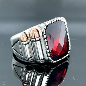 May include: A silver ring with a large red gemstone and three silver bullet-shaped accents. The ring has a detailed design and a polished finish.