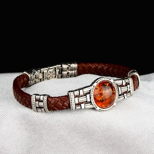 May include: A brown leather bracelet with a silver clasp and a round amber stone in the centre. The stone is surrounded by a silver frame with intricate designs.