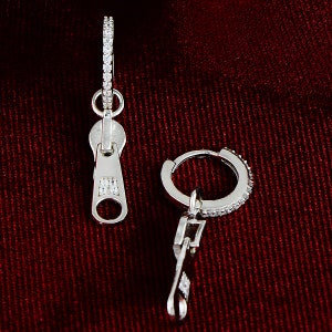May include: A pair of silver earrings with a zipper charm. The earrings are made of a silver hoop with small clear stones around the edge. The zipper charm is also decorated with clear stones.