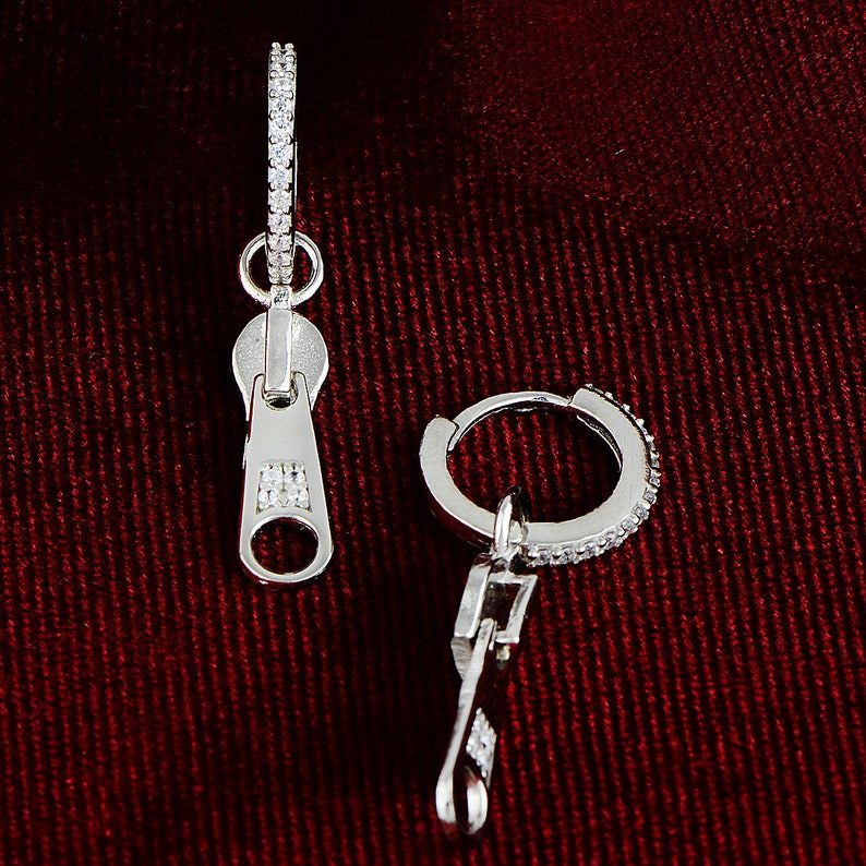 May include: A pair of silver earrings with a zipper charm. The earrings are made of a silver hoop with small clear stones around the edge. The zipper charm is also decorated with clear stones.