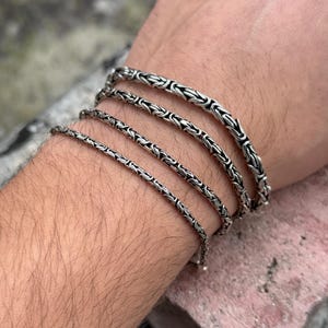 May include: Four silver chain bracelets of varying widths stacked on a person's wrist. The bracelets are made of a tightly woven chain pattern.