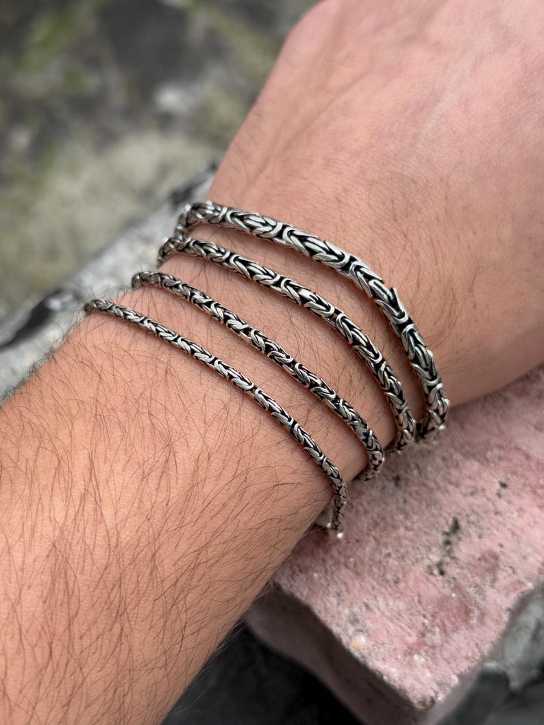May include: Four silver chain bracelets of varying widths stacked on a person's wrist. The bracelets are made of a tightly woven chain pattern.