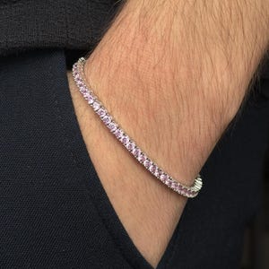 May include: A silver tennis bracelet with a row of pink gemstones. The bracelet is worn on a wrist, with a dark blue shirt in the background. The gemstones are a light pink color.