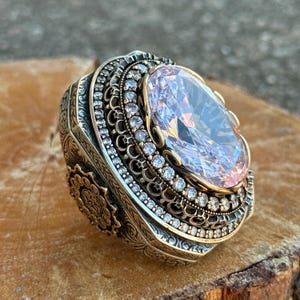 May include: An ornate ring featuring a large, oval-cut, light pink gemstone. The ring has a detailed, engraved band with a gold and silver finish, and is encrusted with small, clear gemstones around the main stone.