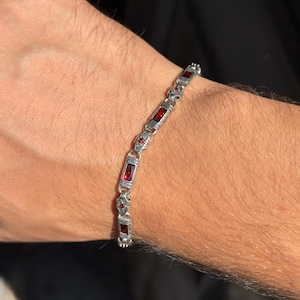 May include: A silver bracelet with rectangular and round red gemstones. The bracelet is made of silver-colored metal and features a series of rectangular red stones interspersed with smaller, round red stones. The bracelet is shown on a person's wrist.