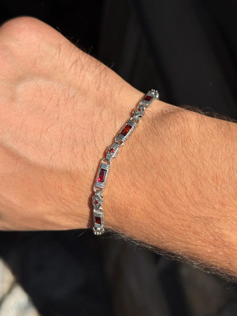 May include: A silver bracelet with rectangular and round red gemstones. The bracelet is made of silver-colored metal and features a series of rectangular red stones interspersed with smaller, round red stones. The bracelet is shown on a person's wrist.