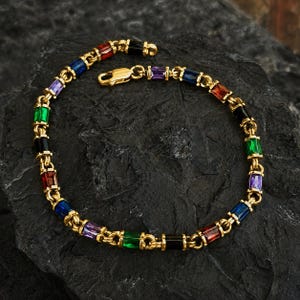 May include: A gold-toned chain bracelet featuring a series of rectangular, multi-colored gemstones in shades of red, blue, purple, green, and black. The bracelet is secured with a lobster claw clasp and displays a repeating pattern of these vibrant stones.