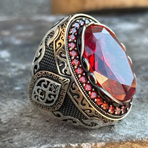 Puede incluir: Un anillo de plata con una piedra preciosa roja ovalada y facetada. El anillo tiene un dise&ntilde;o ornamentado con un borde de piedras preciosas rojas m&aacute;s peque&ntilde;as alrededor de la piedra principal. La banda presenta patrones detallados.