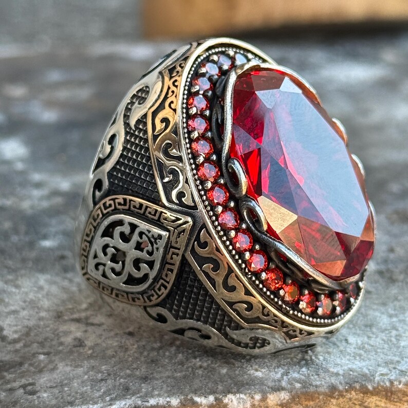 Puede incluir: Un anillo de plata con una piedra preciosa roja ovalada y facetada. El anillo tiene un dise&ntilde;o ornamentado con un borde de piedras preciosas rojas m&aacute;s peque&ntilde;as alrededor de la piedra principal. La banda presenta patrones detallados.