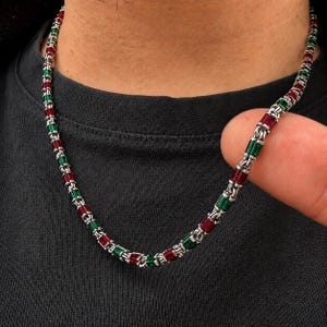 Puede incluir: Un collar de cadena en tono plateado que presenta cuentas rectangulares alternas de color rojo y verde, similares a piedras preciosas. Los eslabones están conectados por pequeños anillos metálicos, creando un patrón continuo de segmentos.