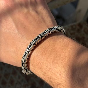 May include: A silver-colored Byzantine chain bracelet. The bracelet is made of interlocking links, creating a textured, woven appearance. The bracelet is worn on a wrist, showcasing its intricate design and metallic sheen.