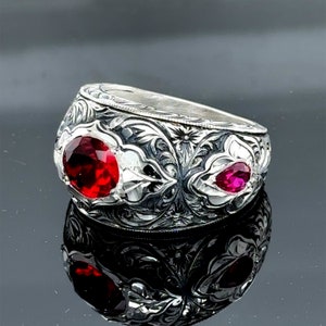 Peut inclure: Une bague en argent avec des motifs floraux complexes et deux pierres pr&eacute;cieuses rouges. La bague est large et a un dessus arrondi.