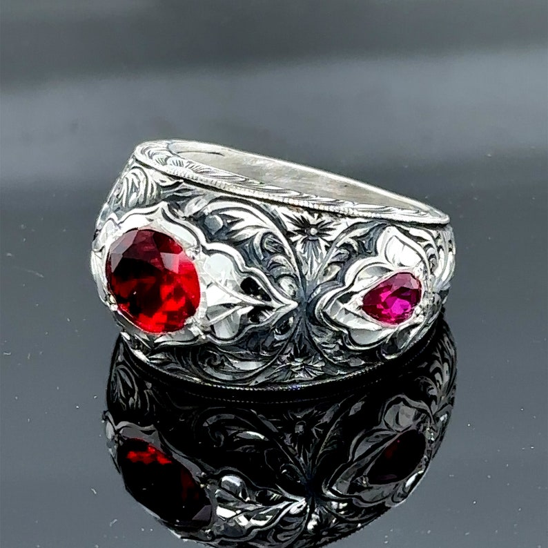 Peut inclure: Une bague en argent avec des motifs floraux complexes et deux pierres pr&eacute;cieuses rouges. La bague est large et a un dessus arrondi.